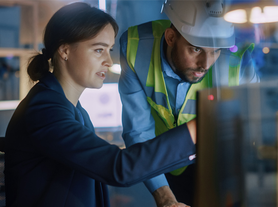 Two colleagues reviewing information on a computer monitor, one wearing a hard hat and safety vest